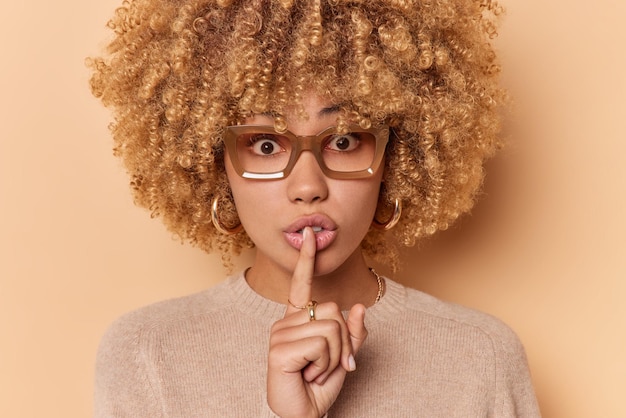 Close up portrait of curly haired woman makes silence gesture keeps index finger over lips has mysterious expression wears spectacles and jumper isolated over beige background. Body language