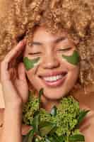 Free photo close up portrait of curly haired woman smiles broadly shows white teeth applies green patches for reducing puffiness undergoes beauty procedures keeps eyes closed. women wellness skin care.