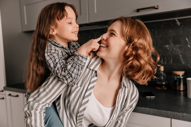 Ritratto del primo piano della giovane mamma dai capelli ricci e della sua piccola figlia allegra in cucina.