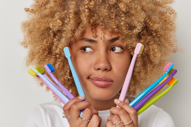 Free photo close up portrait of curly haired young woman holds colorful toothbrushes focused away has serious expression chooses best hygiene product isolated over white background dental care concept