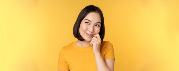 Close up portrait of cute asian girl smiling thinking looking up thoughtful standing in tshirt over yellow background Copy space