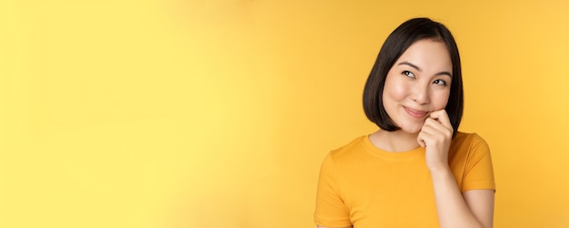 Close up portrait of cute asian girl smiling thinking looking up thoughtful standing in tshirt over ...
