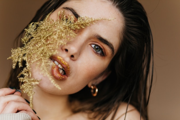 Close-up portrait of cute brunette woman with sparkle lips.  debonair female model holding green plant.