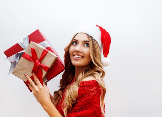 Close up portrait of cute carefree girl with shining wavy blond hairs posing with gift box. Wearing red Santa masquerade hat and sweater.