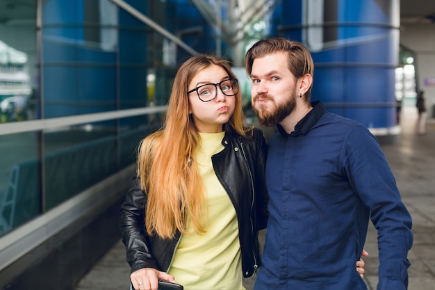 Close-up portrait of cute couple  standing outside in airport. She has long hair, glasses,  yellow sweater, jacket. He wears black shirt, beard. They are  hugging and aping to the camera.