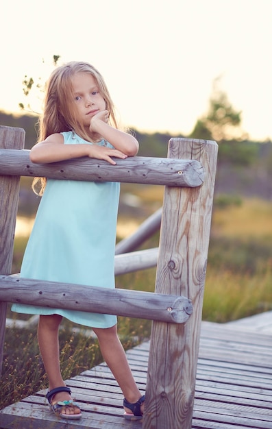 Free photo close-up portrait of a cute little girl, standing outdoors while leaning on a wooden fence.