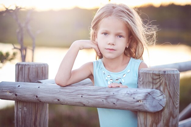 Close-up portrait of a cute little girl, standing outdoors while leaning on a wooden fence.