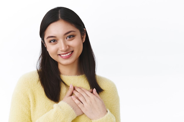 Ritratto ravvicinato ragazza asiatica carina e adorabile premere le mani al cuore, guardando grato e contento, sorridendo mentre riceve un regalo commovente, ringraziando di essere grato, in piedi muro bianco