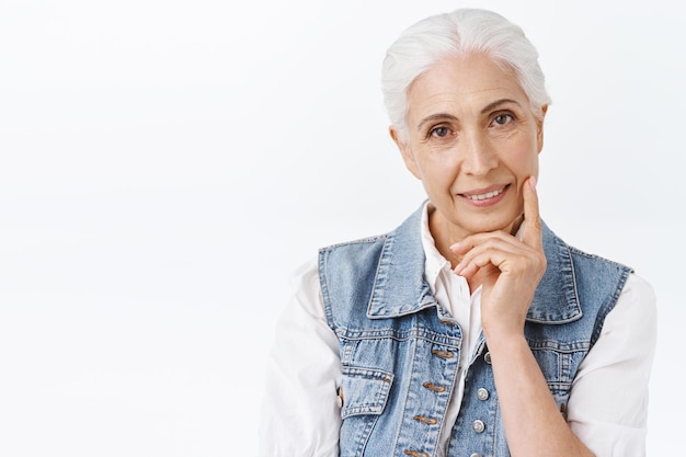 Close-up portrait cute, modern charming senior lady with combed grey hair in denim vest, slightly touching sking and smiling, applying anti-wrinkles cosmetics