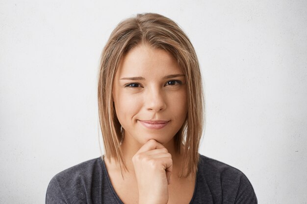 Close-up portrait of cute young female with trendy hairdo and pure skin wearing casual clothes holding hand on cheek looking suspicious posing.