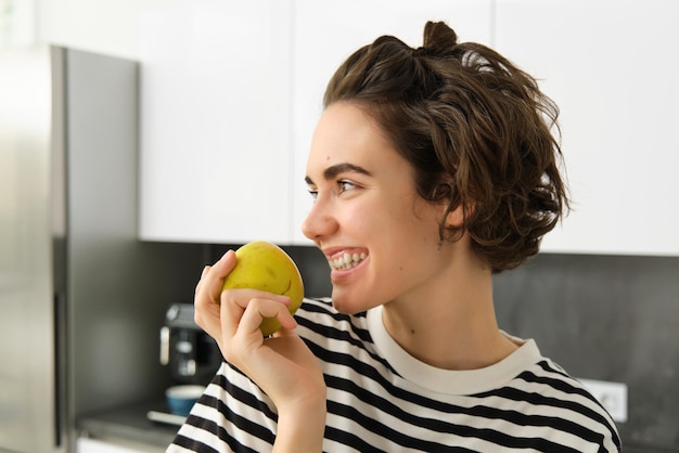 Free photo close up portrait of cute young modern woman eating green apple and looking aside smiling while