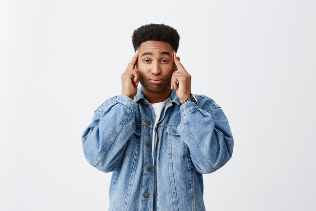 Close up portrait of dark-skinned african men with curly hairstyle in white t-shirt and denim jacket massaging head with fingers, being stressed after hard day on study.