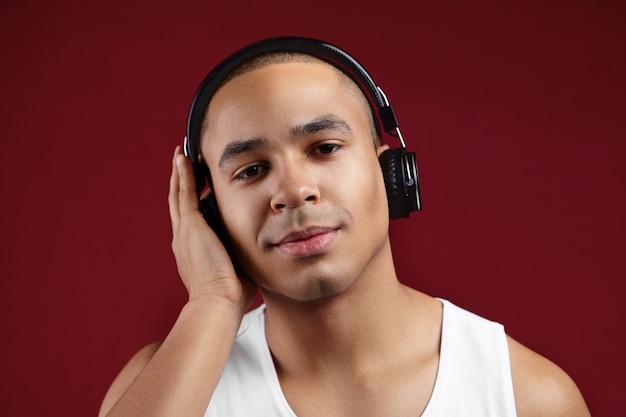 Close up portrait of dark skinned male student wearing black wireless headphones preparing for exams, listening to audio lecture, covering one ear with hand. People, technology, electronic gadgets