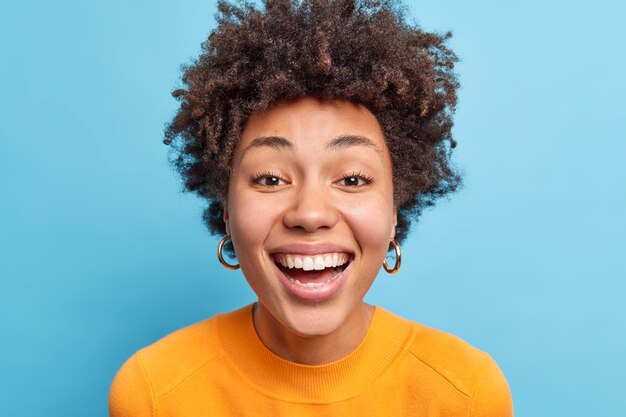 Крупным планом портрет темнокожей женщины с естественными вьющимися волосами, чистой здоровой кожей, улыбкой, широко выражающей счастье, имеет идеальные белые зубы, носит повседневную одежду, изолированную над синей стеной.
