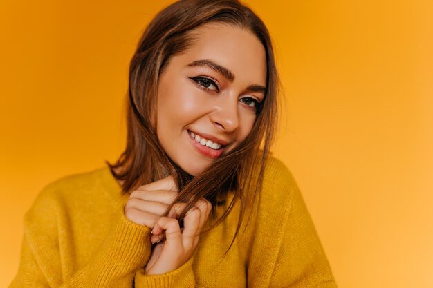 Close-up portrait of debonair female model with short brown hair. Indoor photo of gorgeous caucasian girl smiling on yellow wall.