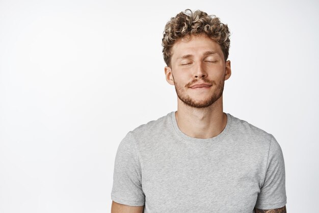 Close up portrait of dreamy blond man close eyes thinking of something daydreaming imaging in his mind standing in gray tshirt over white background