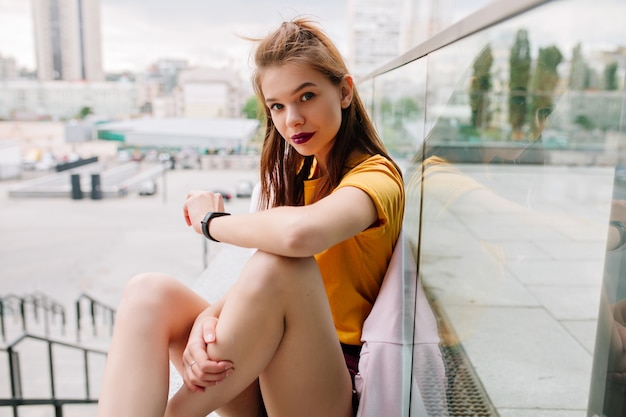 Close-up portrait of dreamy brunette girl sitting on parapet with legs crossed and posing with interest