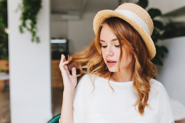 Close-up portrait of dreamy young lady with curly hairstyle and pale skin wearing elegant straw hat decorated with white ribbon