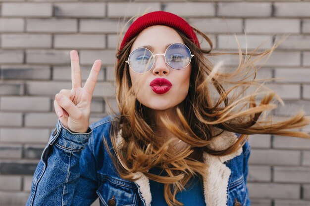 Close-up portrait of ecstatic white girl spending spring day outdoor. Photo of inspired blonde woman wears denim jacket and red hat posing with kissing face expression.