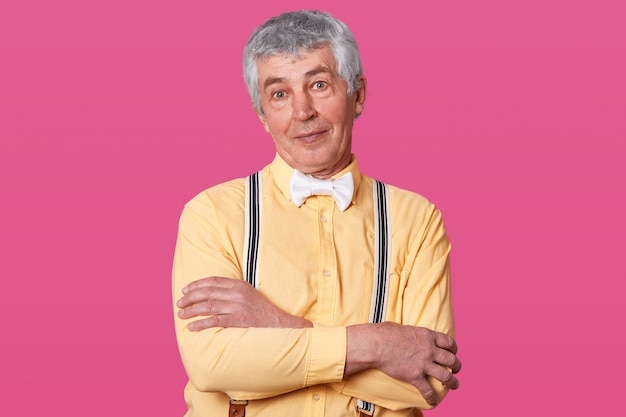 Close up portrait of eldery man with yellow shirt and white bow tie, looking directly at camera, keeps hands folded, free spase for your advertisment or promotion, isolated on rose studio.