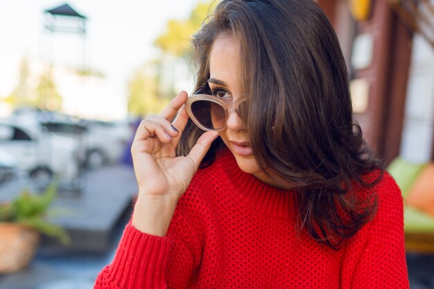 Close up portrait of elegant romantic woman with brunette wavy hairs with stylish retro sunglasses and wed knitted sweater. Female chilling in modern cafe in the morning and drink coffee.