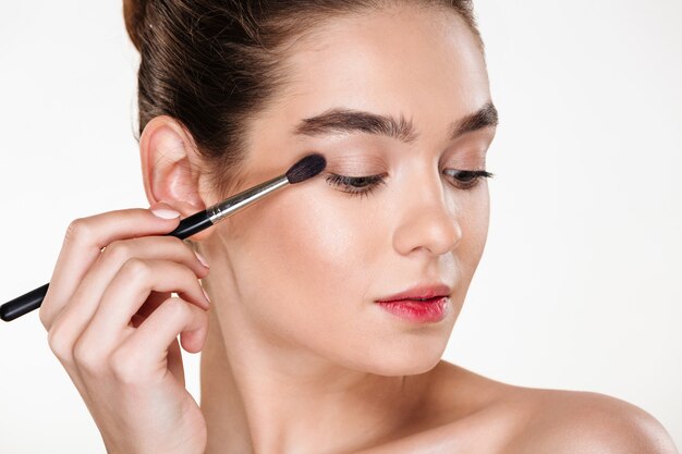 Close up portrait of elegant woman with clean shining skin applying eye shadow using brush