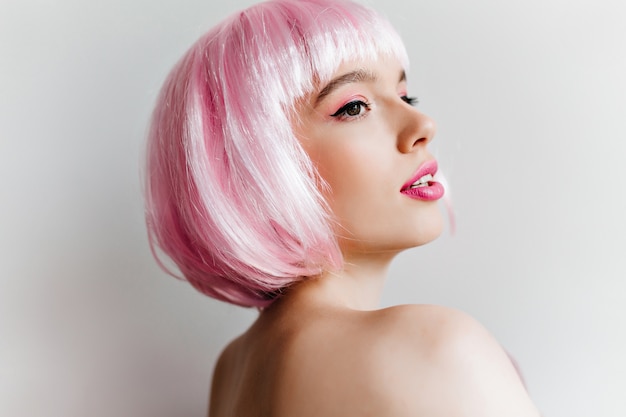 Крупным планом портрет элегантной молодой женщины в розовом парике, глядя с интересом. Удивительная кавказская девушка с короткими прямыми волосами позирует на светлой стене.