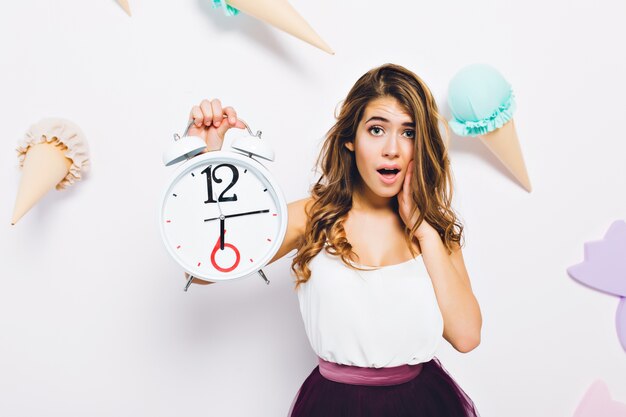 Close-up portrait of elegant young woman in stylish dress, posing with white clock on decorated wall. Long-haired curly girl with unhappy face expression standing in front of wall with ice cream