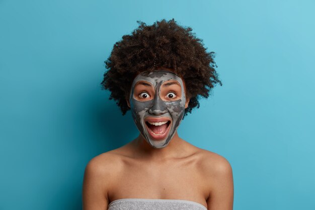 Free photo close up portrait of emotional excited dark skinned woman with afro hairstyle exclaims positively, relaxes with cosmetic mask on face, being late for date, stands wrapped in towel. spa, health care