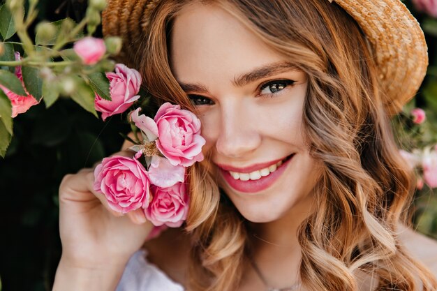 Портрет крупным планом очаровательной девушки с блестящими глазами, позирующей с цветком. Эффектная блондинка в шляпе, держа розовую розу и улыбается.