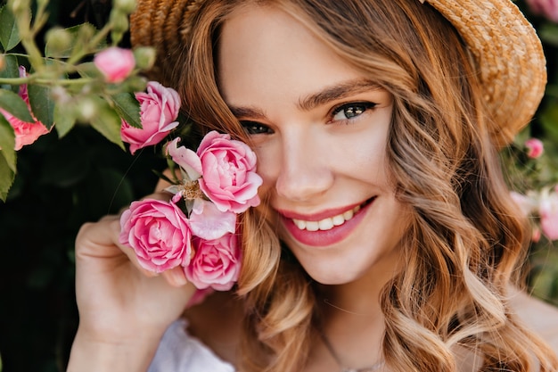 Close-up portrait of enchanting girl with shiny eyes posing with flower. Spectacular blonde woman in hat holding pink rose and smiling.