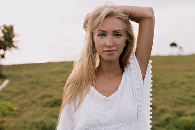 Close up portrait of european woman with long hair dressed in white clothes posing at camera on background of beach with green plants