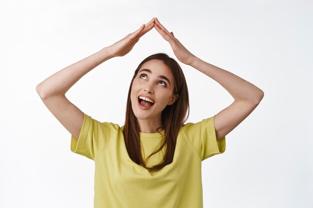 Close up portrait of excited smiling girl building roof house with hands above head, feel secured, looking happy at rooftop, standing over white background