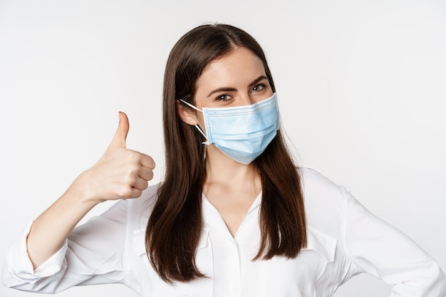 Close up portrait, face of woman in medical mask smiling, showing thumbs up, standing over white background