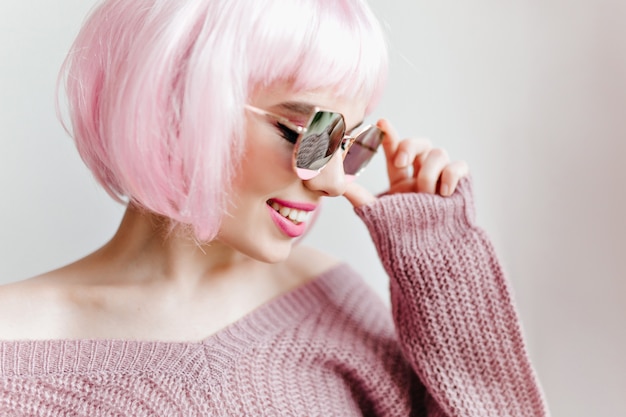 Портрет крупным планом очаровательной коротко стриженной дамы в очках, позирующей на светлой стене. женская модель в модном розовом перуке.
