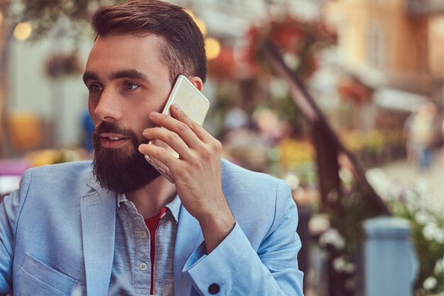 Close-up portrait of a fashionable bearded businessman with a stylish haircut, speaking by phone, drinks a glass of a cool juice, sitting in a cafe outdoors.