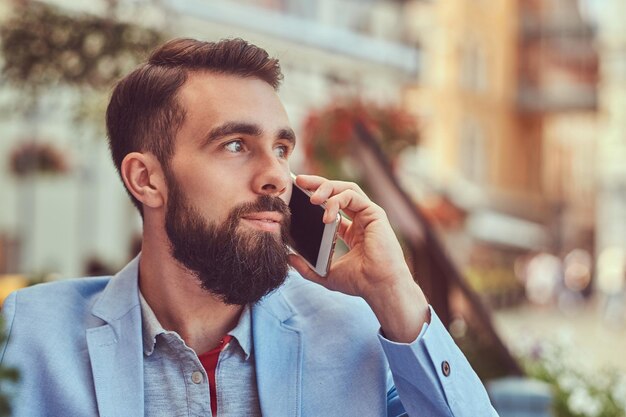 Close-up portrait of a fashionable bearded businessman with a stylish haircut, speaking by phone, drinks a glass of a cool juice, sitting in a cafe outdoors.