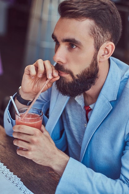세련된 머리를 한 세련된 수염 난 남성의 클로즈업 초상화는 야외 카페에 앉아 주스 한 잔을 마신다.