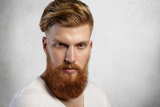 Close up portrait of fashionable redhead hipster man with fuzzy beard and trendy haircut wearing white t-shirt while posing isolated against wall   with serious or angry expression