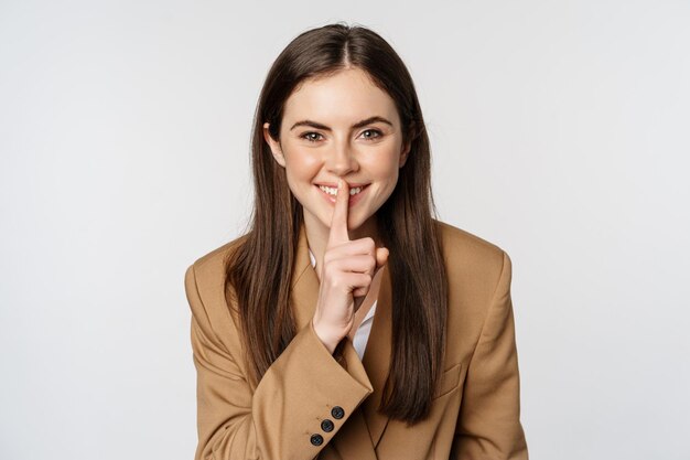 Close up portrait of female entrepreneur smiling shushing making shh hush sign press finger to lips ...