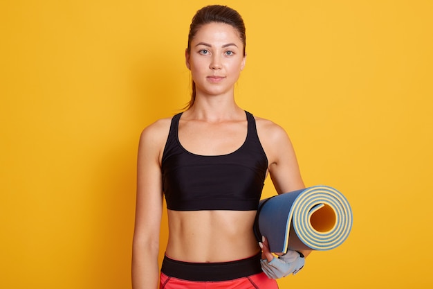 Close up portrait of fitness woman ready for workout, slim girl standing against yellow studio wall and holding yoga mat