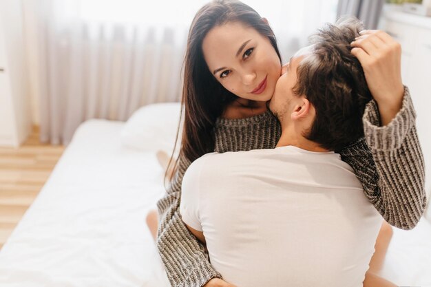 Close-up portrait from back of brunette guy embracing pretty european girl in bedroom. Indoor photo of smiling beautiful lady in woolen shirt touching husband's hair.