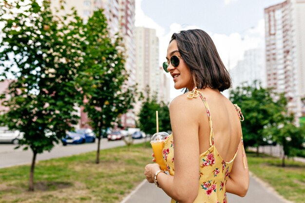 Close up portrait from back of lovely girl with short hairstyle in summer dress and sunglasses walking in the city