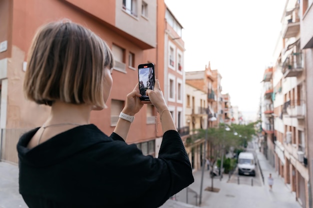 Close up portrait from back of modern stylish girl with short hairstyle is making photo of city view on smartphone in summer evening Debonair shorthaired woman standing on the street with phone