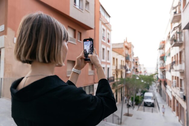 短い髪型のモダンでスタイリッシュな女の子の後ろからの肖像画をクローズアップは、夏の夜にスマートフォンで街の景色の写真を作っています電話で通りに立っているデボネアの短い髪の女性