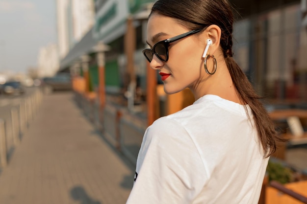 Close up portrait from back of stylish girl with dark hair and red lips is wearing dark sunglasses and golden earrings