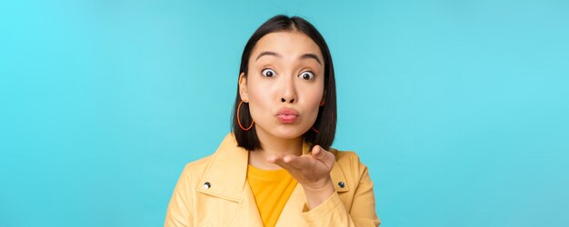 Close up portrait of funny asian girl sending air kiss blowing at camera with popped eyes standing over blue background