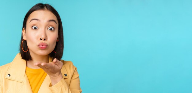 Close up portrait of funny asian girl sending air kiss blowing at camera with popped eyes standing over blue background