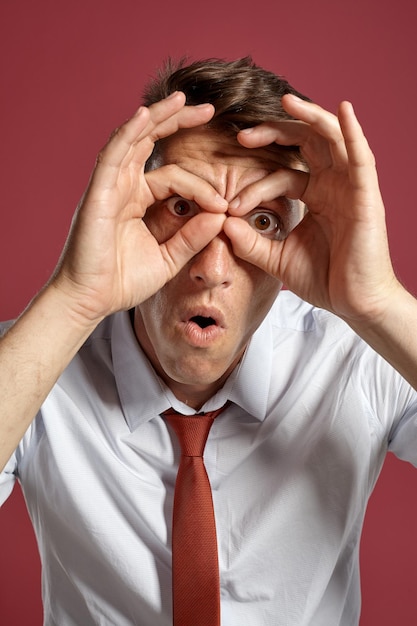 Free photo close-up portrait of a funny brunet person with brown eyes, wearing in a white shirt and a terracotta tie. he is fooling around while posing in a studio against a red background. concept of gesticulat