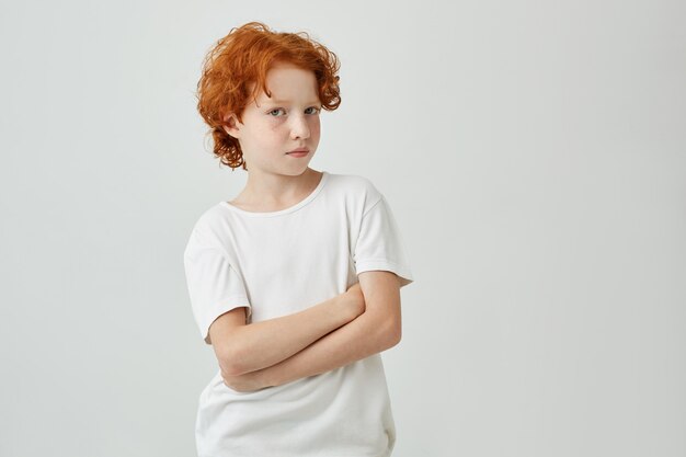 Закройте вверх по портрету смешного мальчика имбиря с веснушками в белой футболке смотря с расслабленным выражением и пересеченными руками.